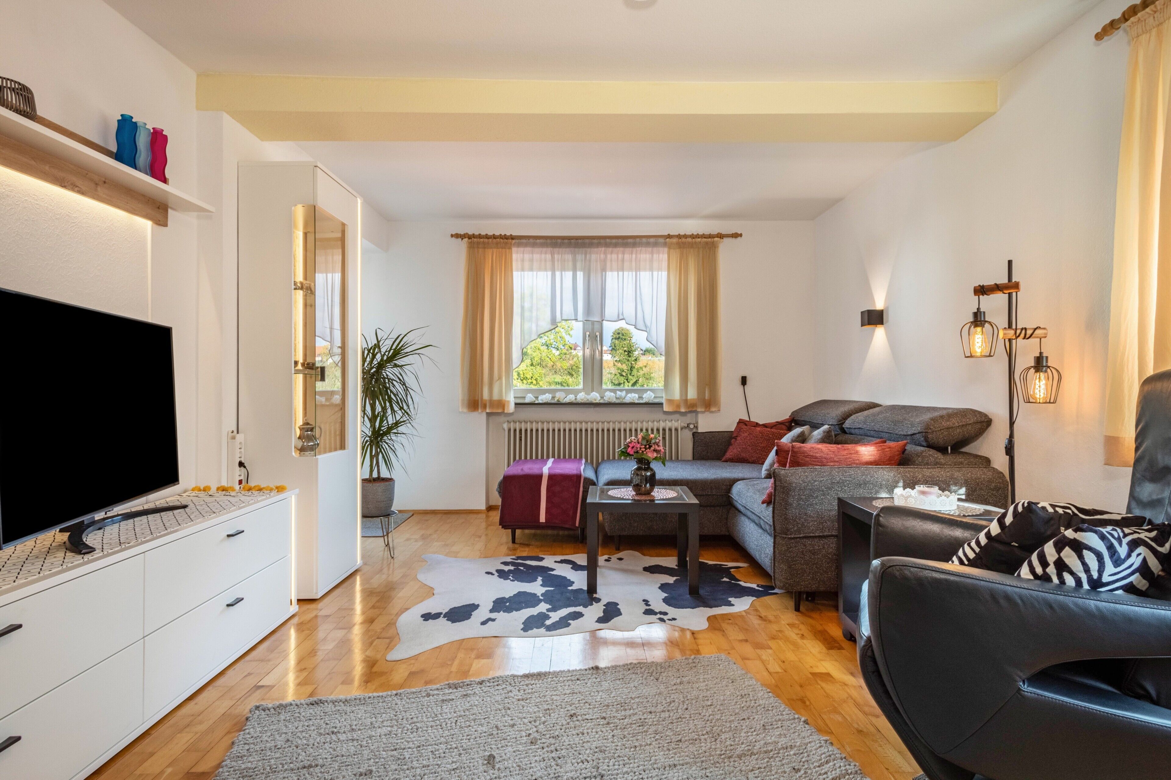 A modern living room with a flat-screen TV, a sofa, a coffee table, and a large window with curtains.