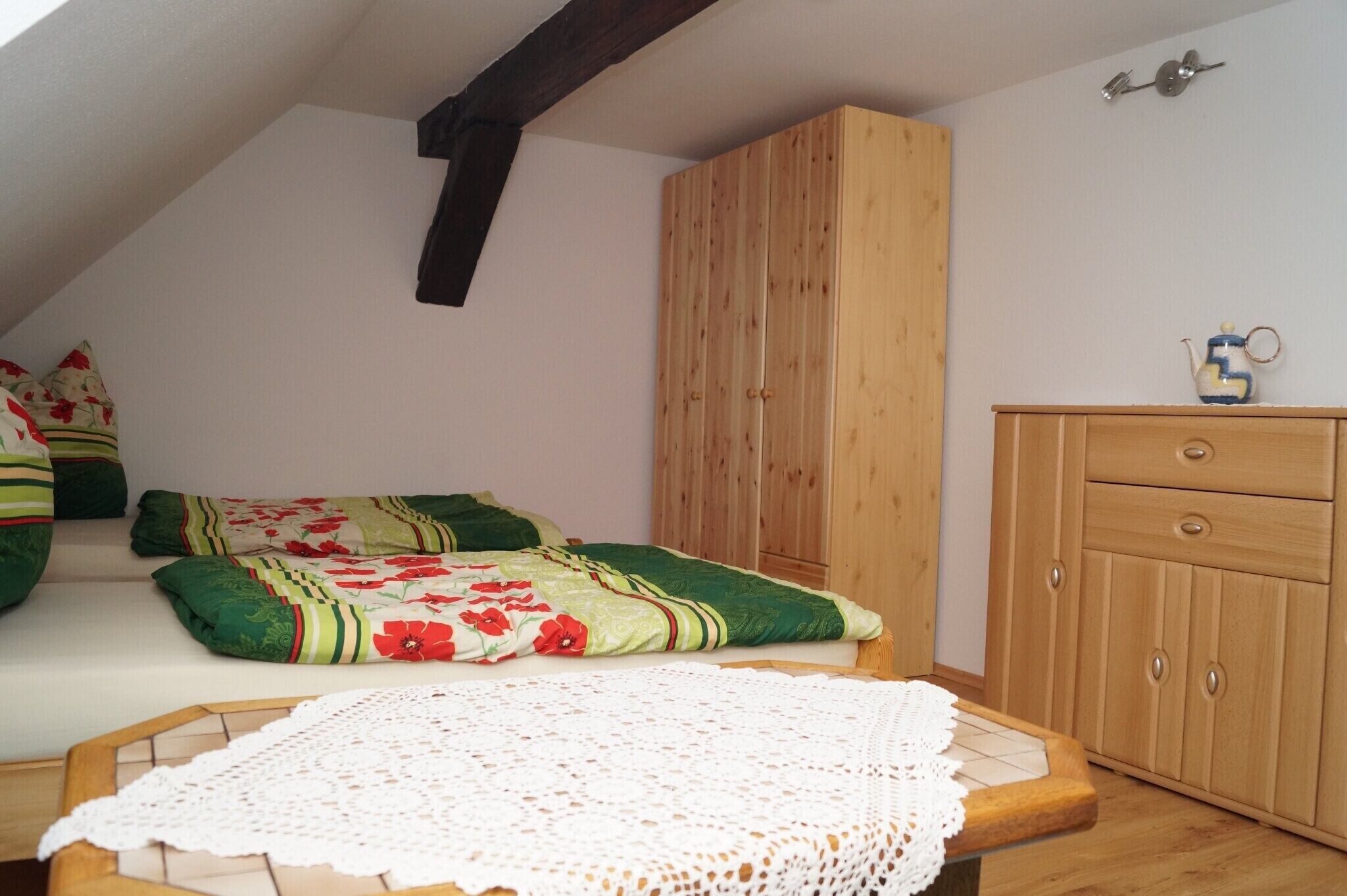 A room with a sloped ceiling, a wooden wardrobe, a bed with floral bedding, and a small table with a white lace tablecloth.