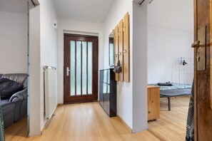 Een smalle gang met een houten vloer, een radiator, een deur met geëtste glaspanelen, een bed met matras en kussens, en een klein houten kastje.