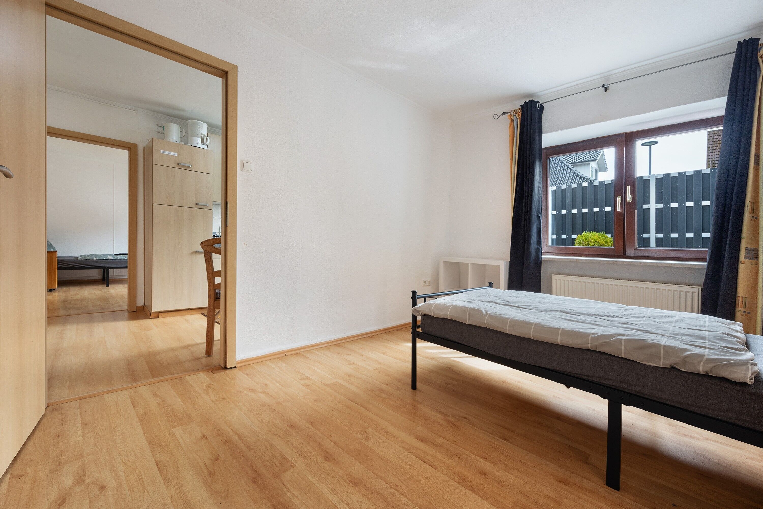 Une chambre à coucher avec un lit, un plancher en bois, une fenêtre avec des rideaux et une vue sur une clôture.
