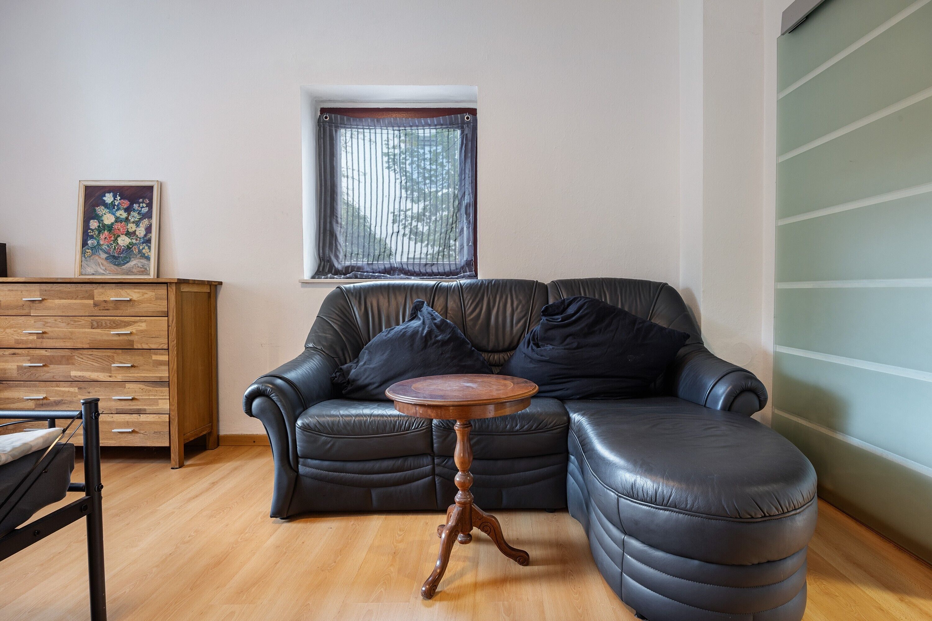 Un salon avec un canapé en cuir, une table d’appoint en bois, une commode en bois et une fenêtre avec des stores.