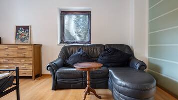 Un salon avec un canapé en cuir, une table d’appoint en bois, une commode en bois et une fenêtre avec des stores.