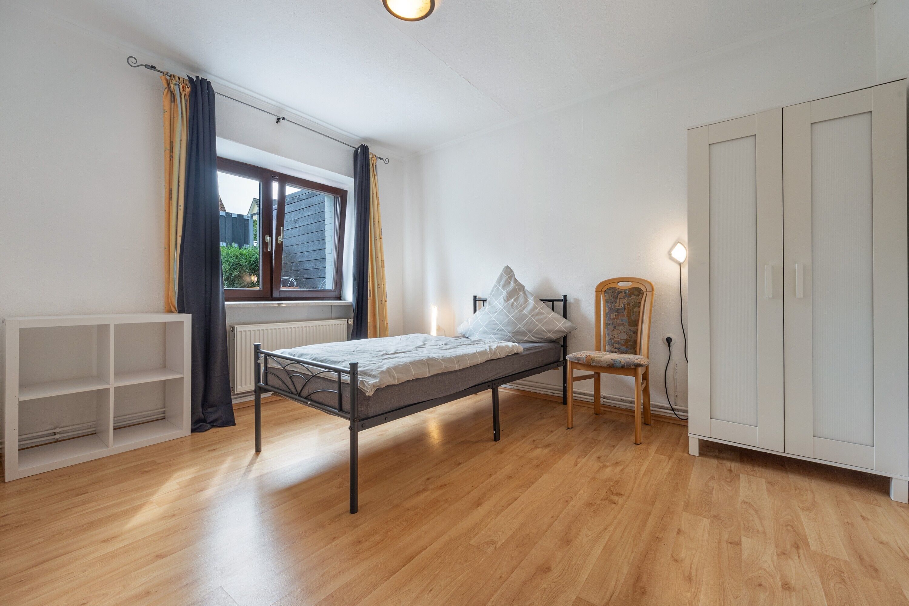 A bedroom with a single bed, a chair, a wardrobe, and a window with curtains.