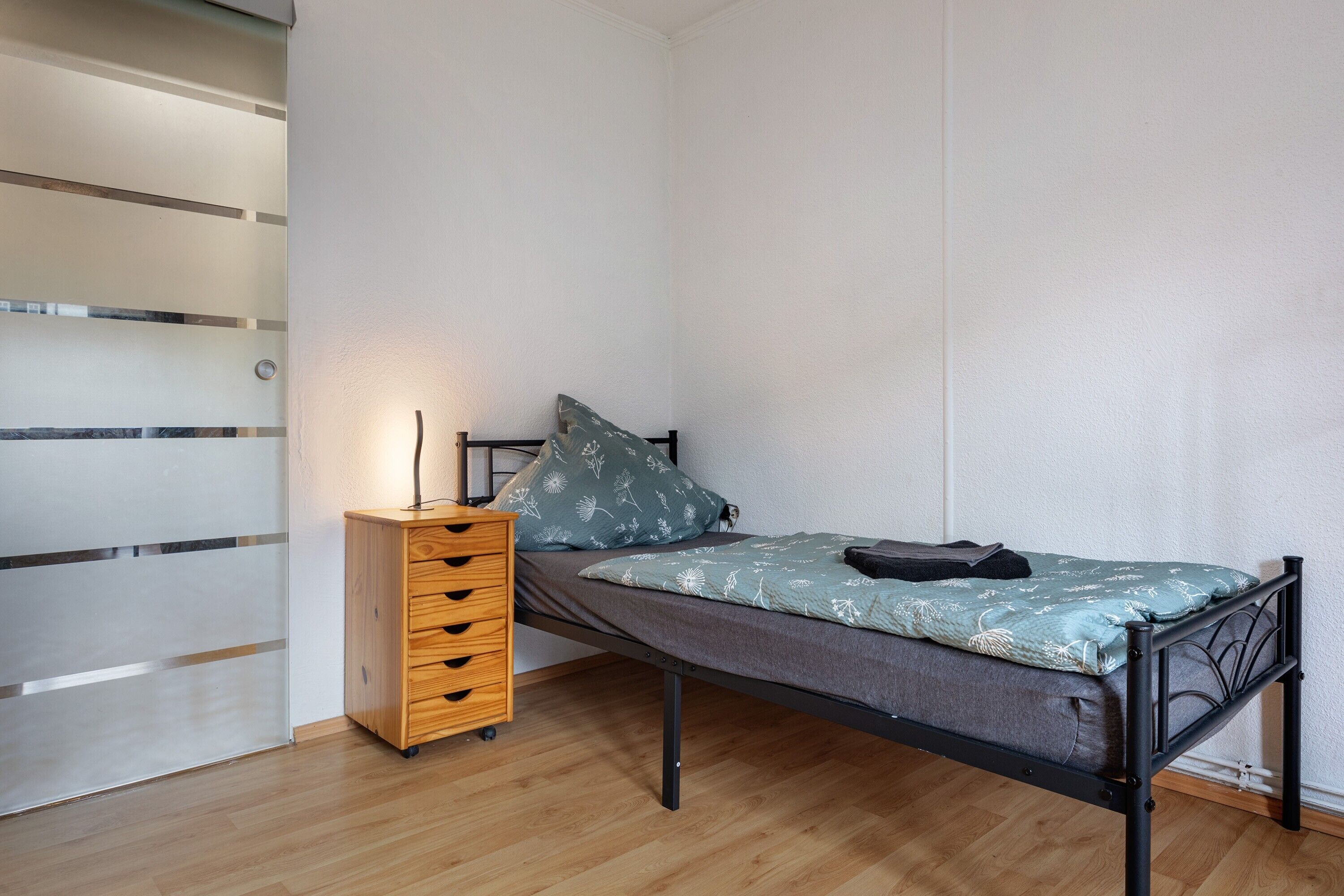 A single bed with a metal frame, a wooden dresser with drawers, and a wall with a frosted panel.