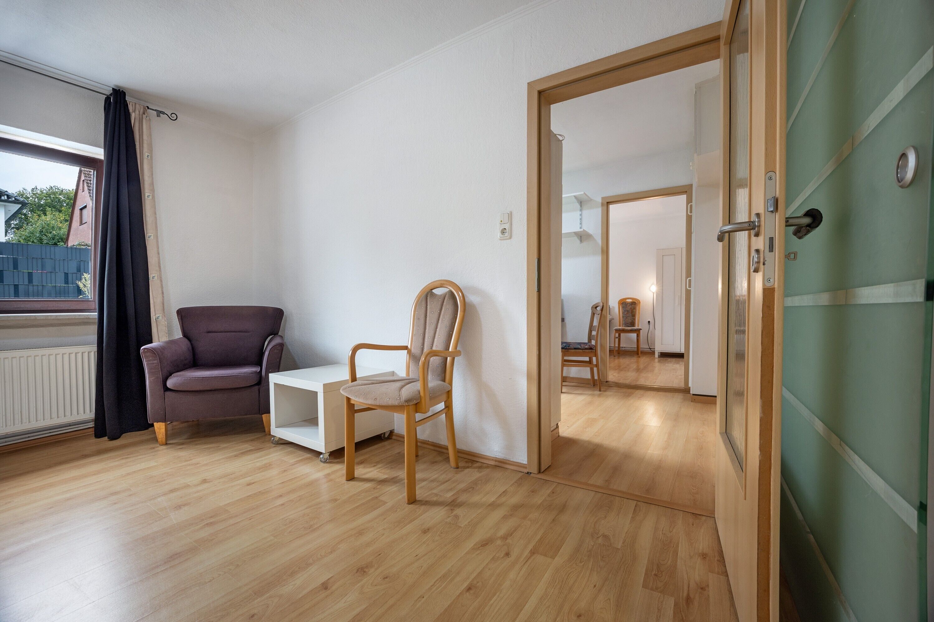 Une pièce avec un plancher en bois, un fauteuil violet, une chaise en bois, une petite table d’appoint blanche et une porte qui donne sur une autre pièce.