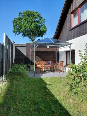 Een overdekt terrasgedeelte met een tafel en stoelen, omgeven door een houten omheining en een boom.