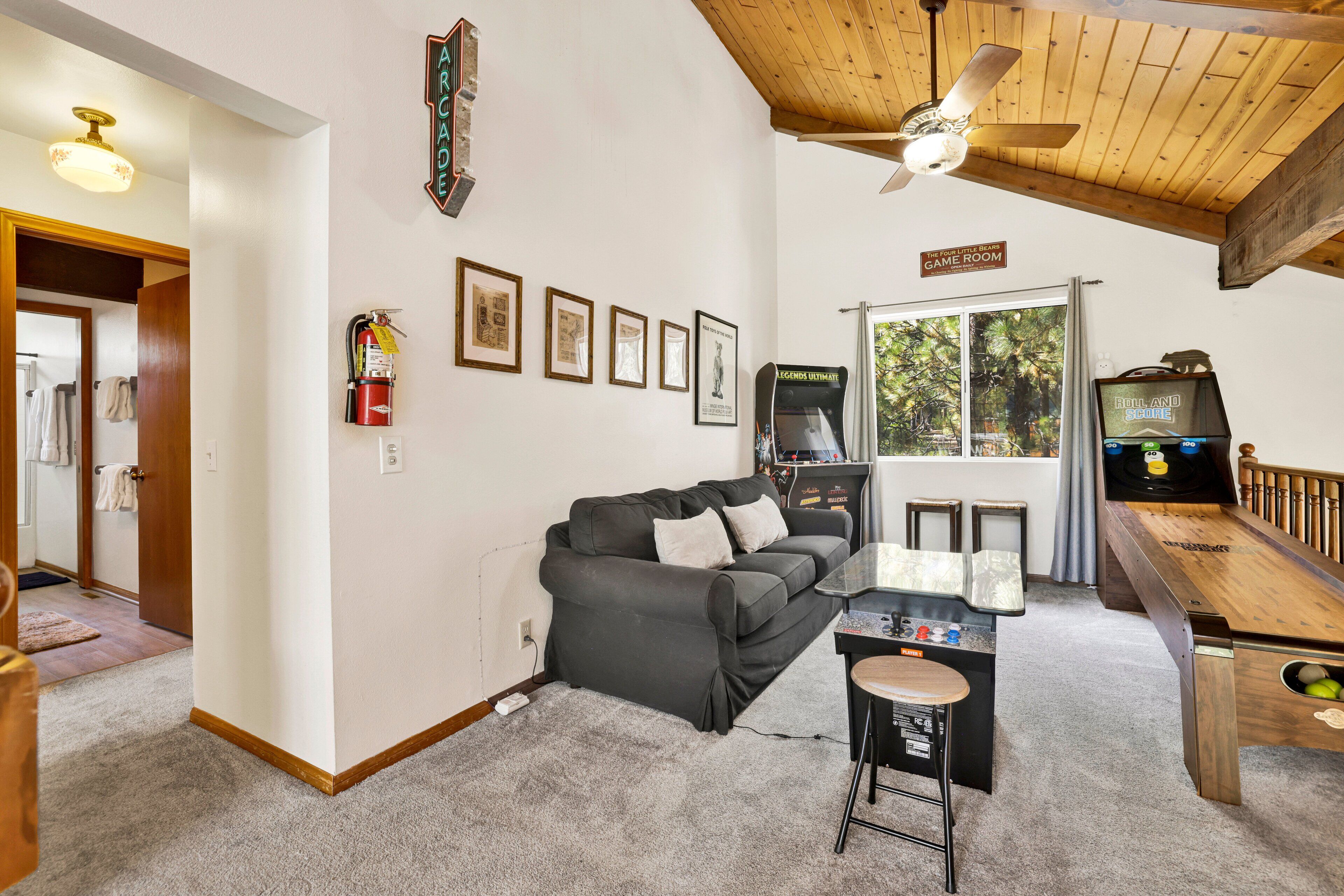 A cozy living room with a gray couch, a wooden coffee table, and a ceiling fan. There are framed pictures on the wall and a fire extinguisher mounted on the wall.