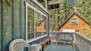 A wooden deck with wicker chairs and a bench, overlooking a forested area and a cabin.