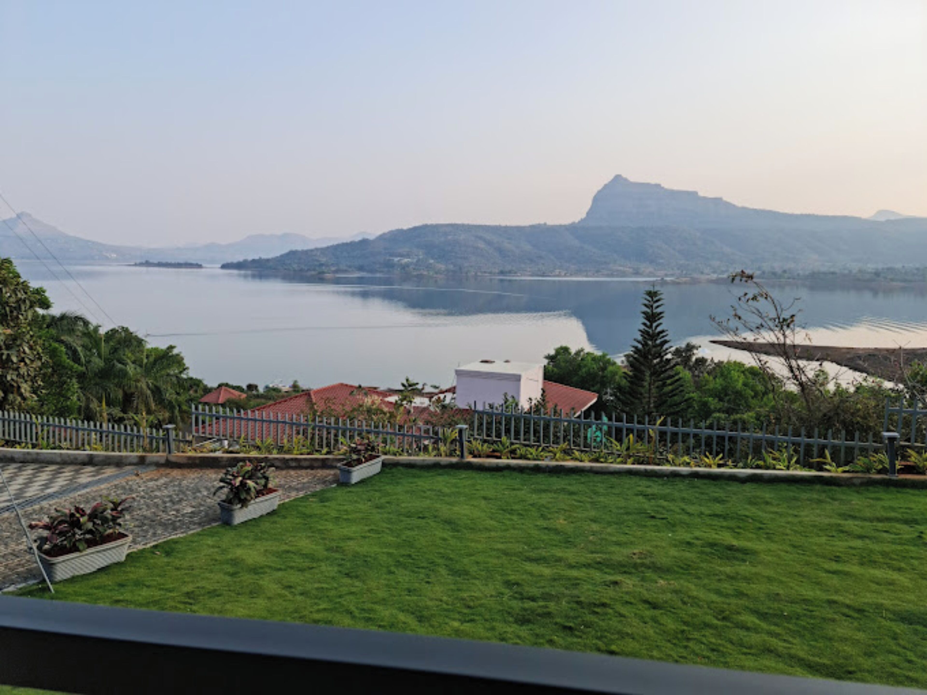 A view of a lake with mountains in the background, a fenced garden area with grass and potted plants, and a white building in the foreground.