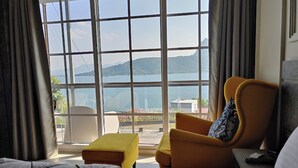 A hotel room with a large window offering a view of a lake and mountains. There is a bed, a yellow armchair, and a small table with a remote control. - Jumbo Paradise (Mawal)