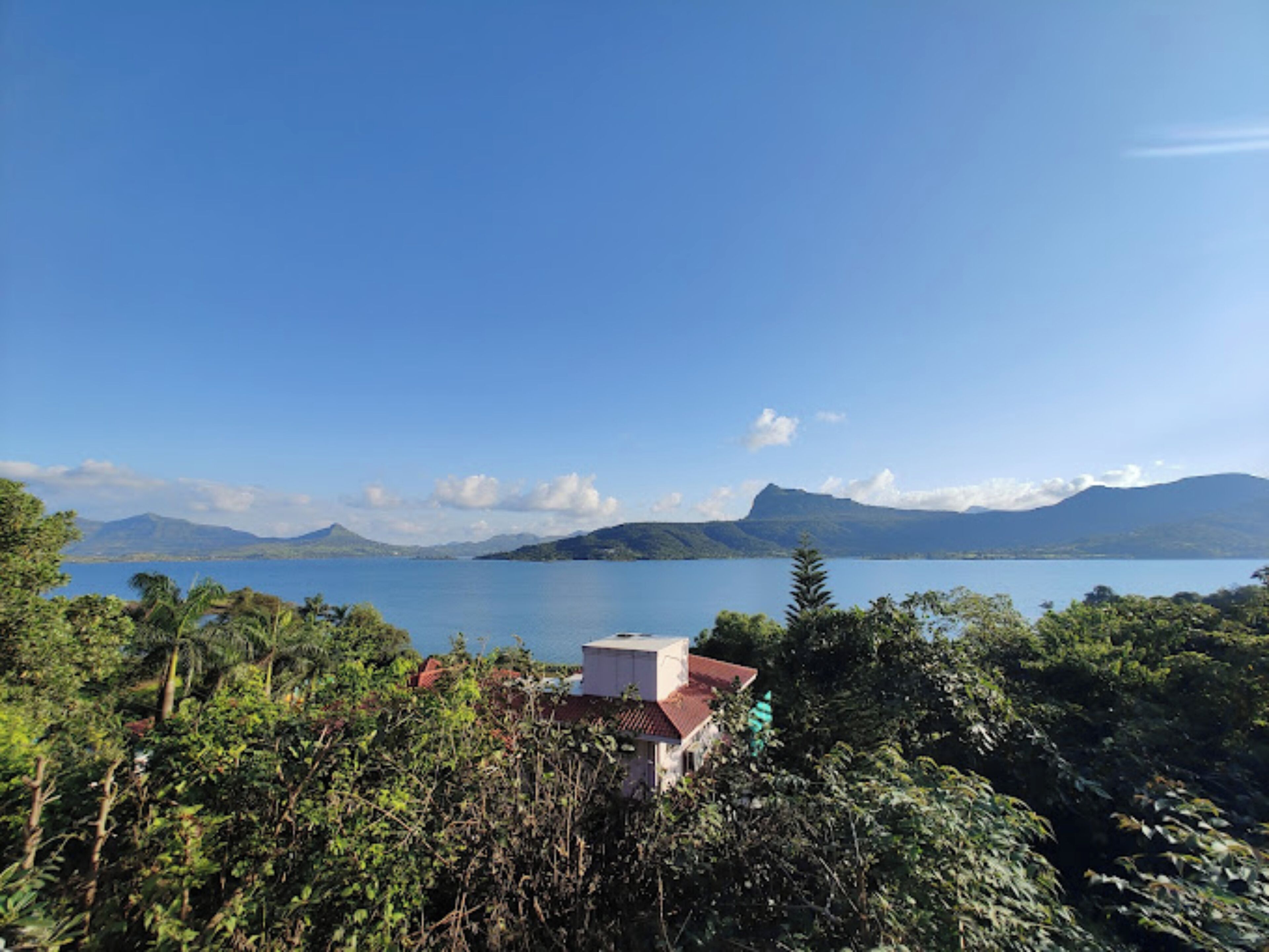 A house with a red roof surrounded by dense greenery, overlooking a calm lake with mountains in the distance.