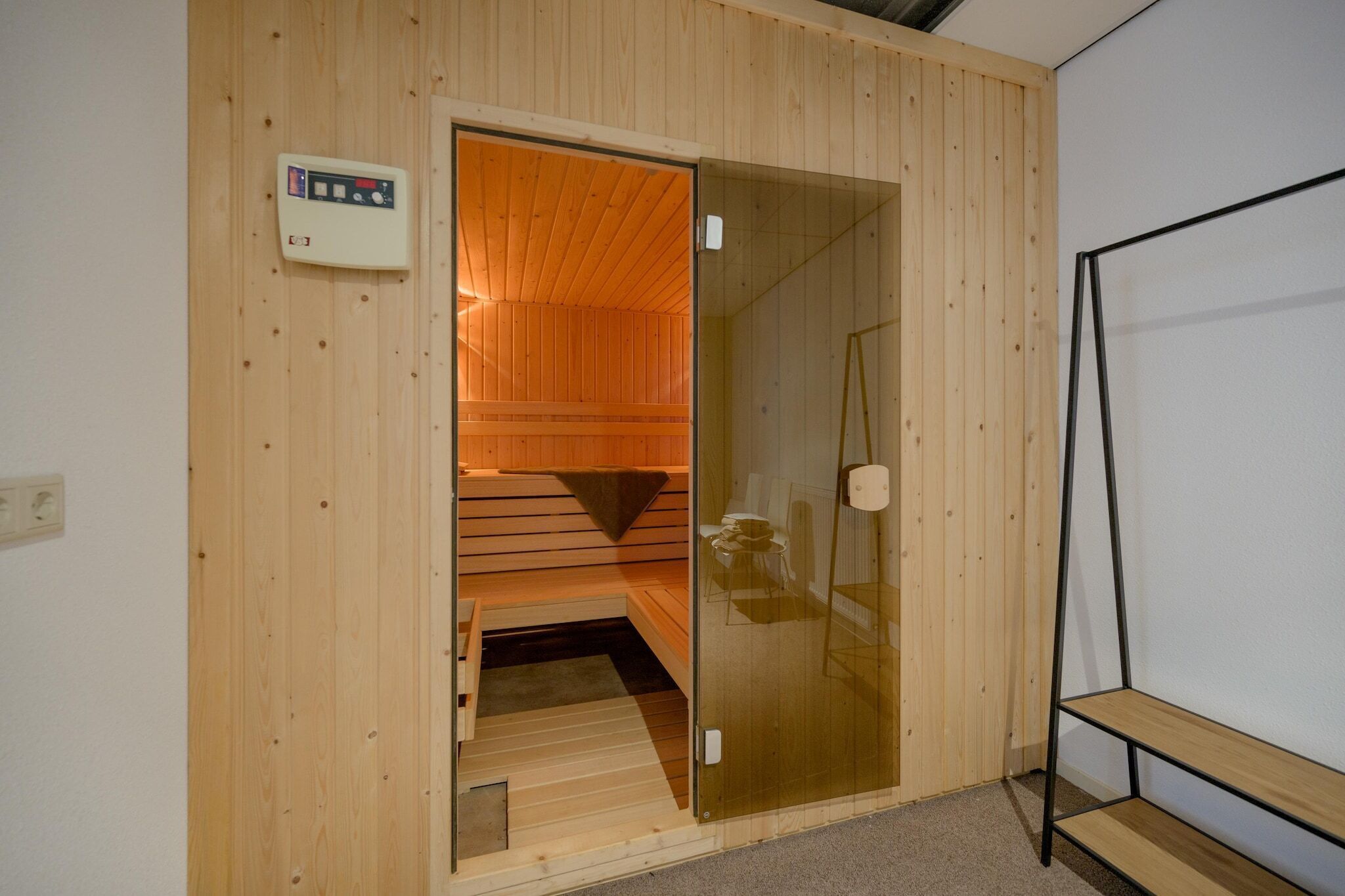 A sauna with wooden walls and a glass door, a control panel, and a shelf.