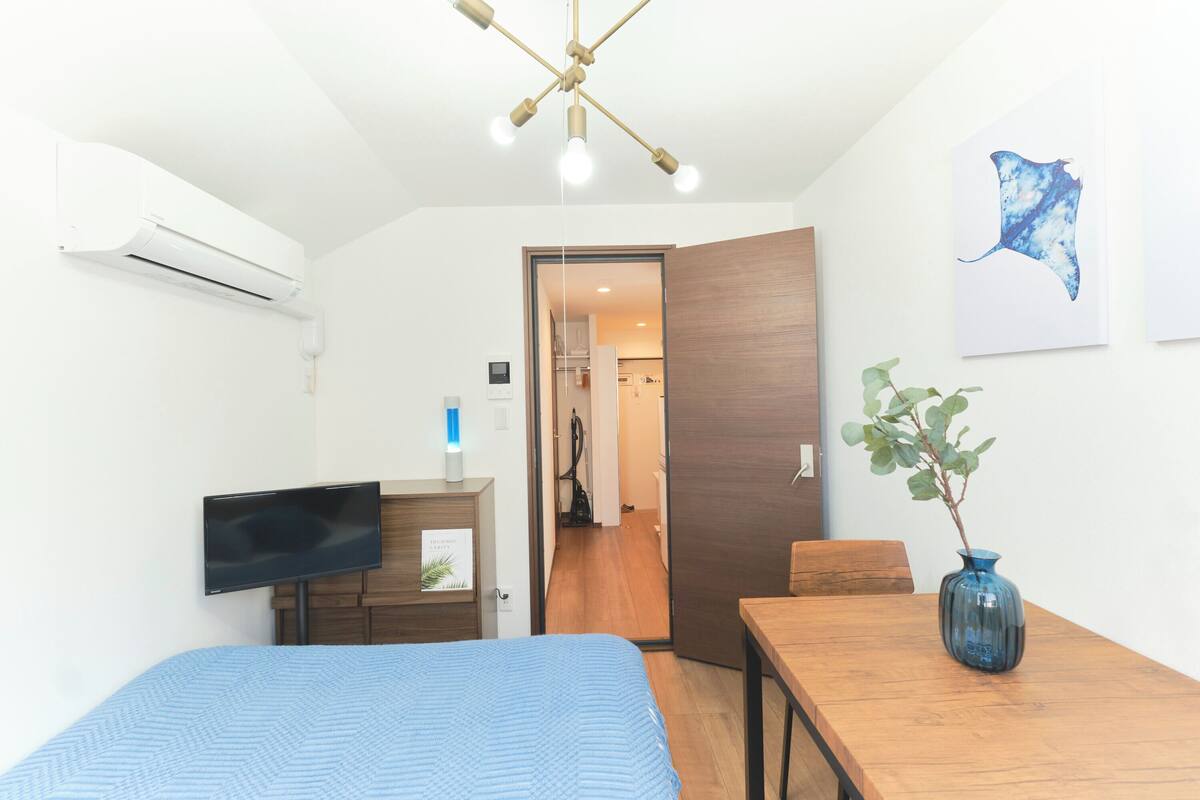 A bedroom with a bed, a TV, a desk, a chair, a wooden table, a vase with a plant, and a ceiling light fixture.