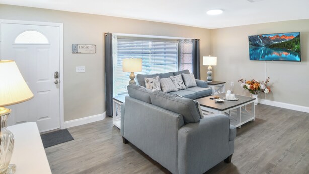 A living room with a grey sofa, a coffee table, a lamp, a painting on the wall, and a door labeled "HOME". - The Blooming Sanctuary - 3br with pool and EV-Charger (Rotonda West)