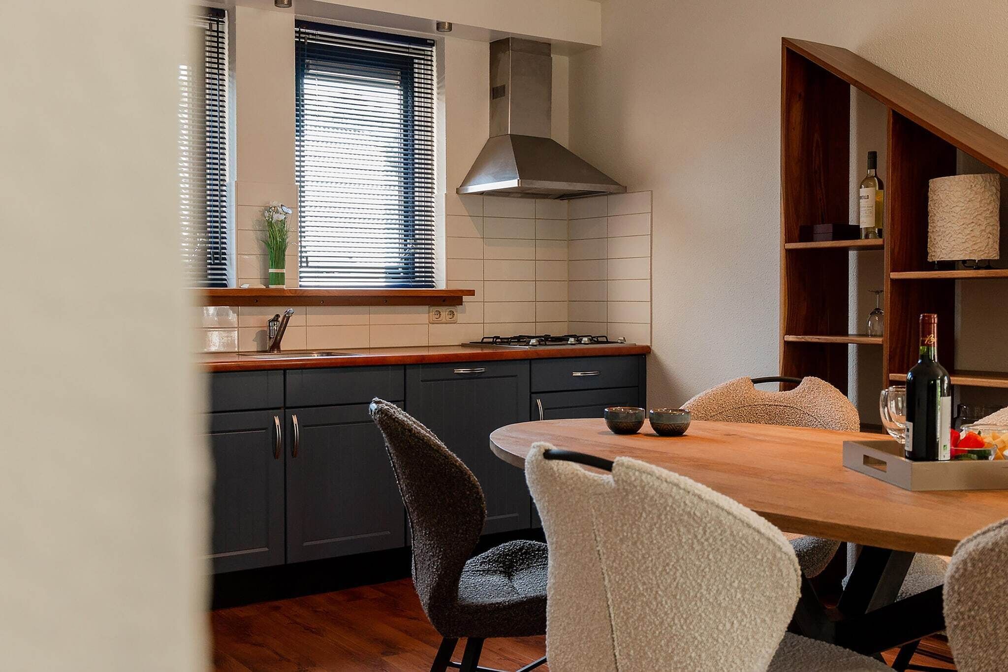 Une cuisine moderne avec des meubles sombres, un plan de travail en bois, une cuisinière à gaz et une hotte intégrée. On y trouve une table à manger avec des chaises et une bouteille de vin sur l’étagère.