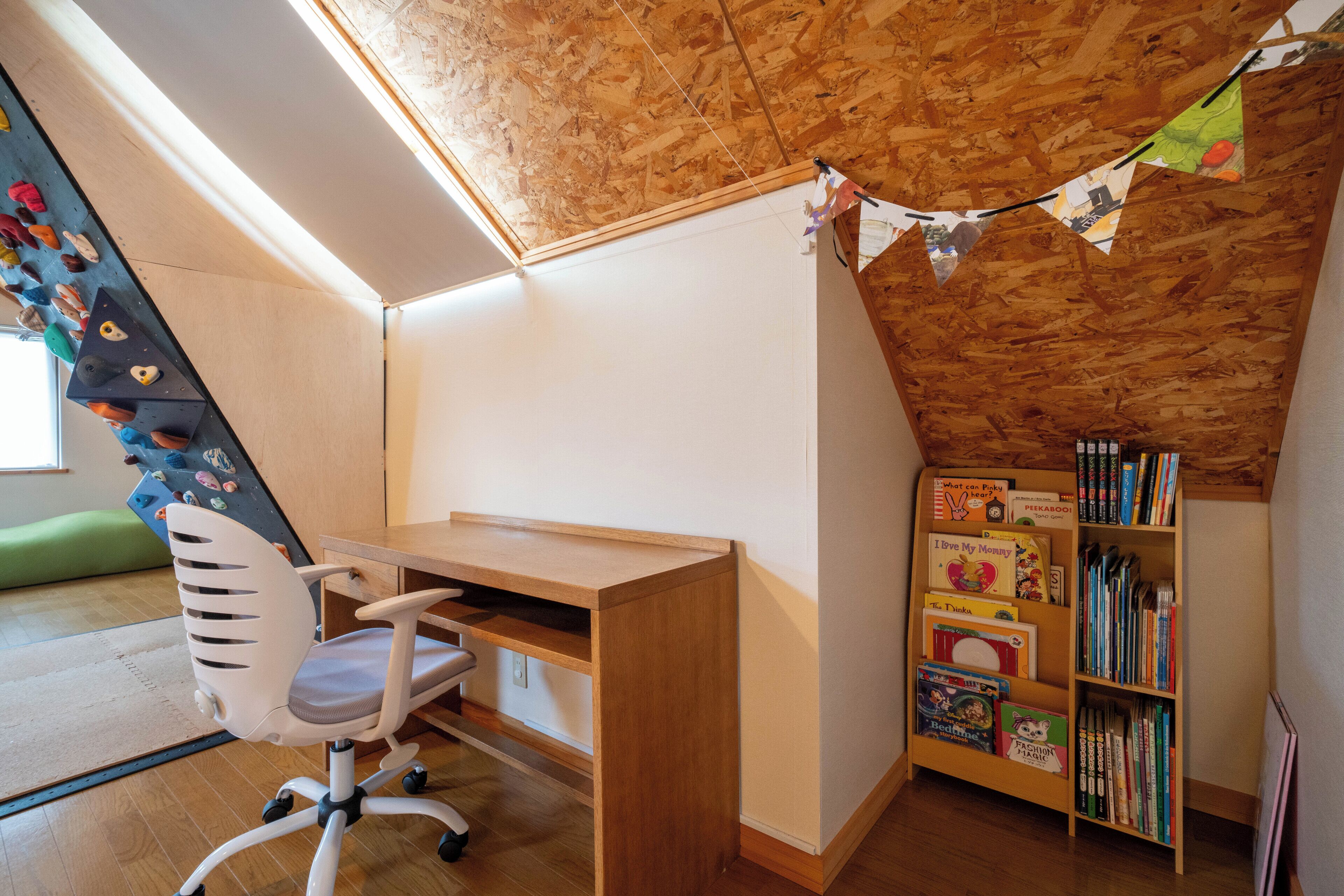 A room with a sloped ceiling, a wooden desk, a white chair, a bookshelf, and a climbing wall.