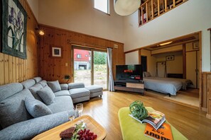 A modern living room with a grey sectional sofa, a wooden coffee table with a magazine and grapes, a flat-screen TV, and a bed in the adjacent room. - RONIN LODGE (Furano)