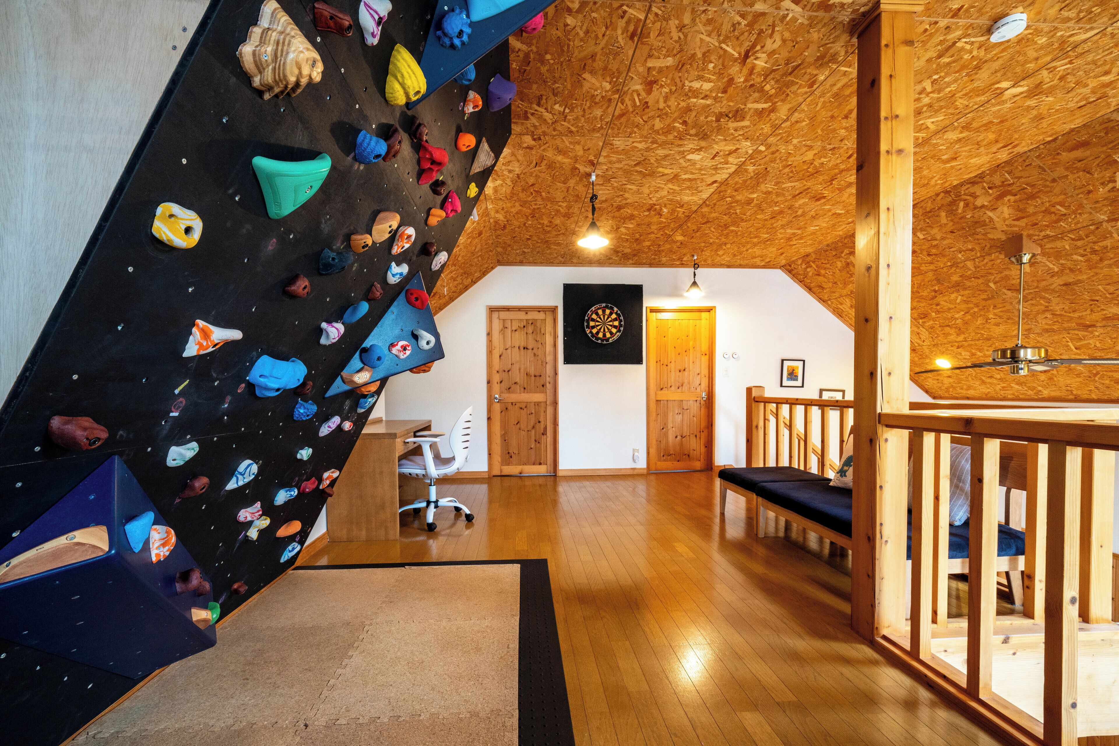 A climbing wall with various holds, wooden floors, a ceiling fan, and a dartboard on the wall.
