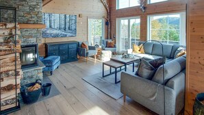 Ein gemütliches Wohnzimmer mit Kamin aus Stein, Holzbalken und großen Fenstern mit Blick ins Freie.