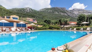 Piscina con sillas de descanso y sombrillas, rodeada de montañas y follaje.