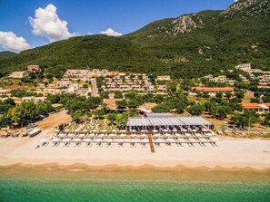 A coastal town with a beach area featuring rows of sun loungers and umbrellas, surrounded by residential buildings and a mountainous backdrop.