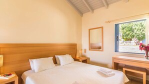 Habitación de hotel con cabecera de madera, una cama grande con sábanas blancas, una mesita de noche con lámpara, un escritorio con un jarrón de flores y una ventana con vistas a zonas verdes.