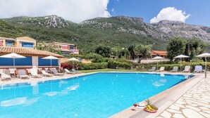 A swimming pool with lounge chairs and umbrellas, surrounded by buildings and mountains. - Akti Barbati Houses by Konnect - Two-Bedroom Maisonette (Corfu)
