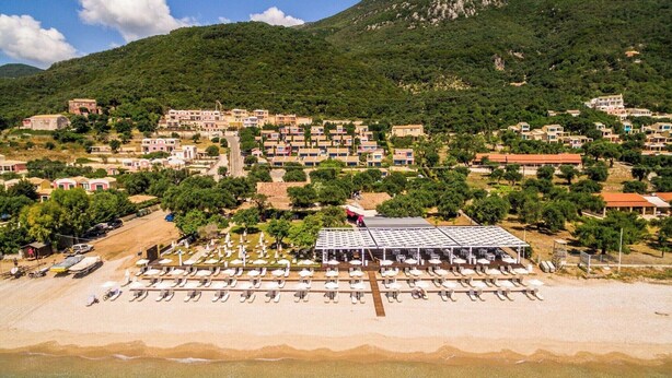 A coastal town with a beach area featuring rows of sun loungers and umbrellas, surrounded by residential buildings and a mountainous backdrop. - Akti Barbati Houses by Konnect - Two-Bedroom Maisonette (Corfu)