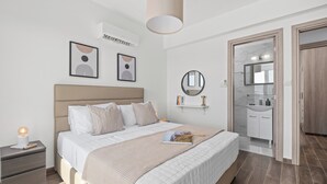 A modern bedroom with a large bed, bedside tables, a bedside lamp, a wall-mounted air conditioner, a ceiling light, a framed artwork, a round mirror, a bathroom with a sink and mirror, and a wooden door.