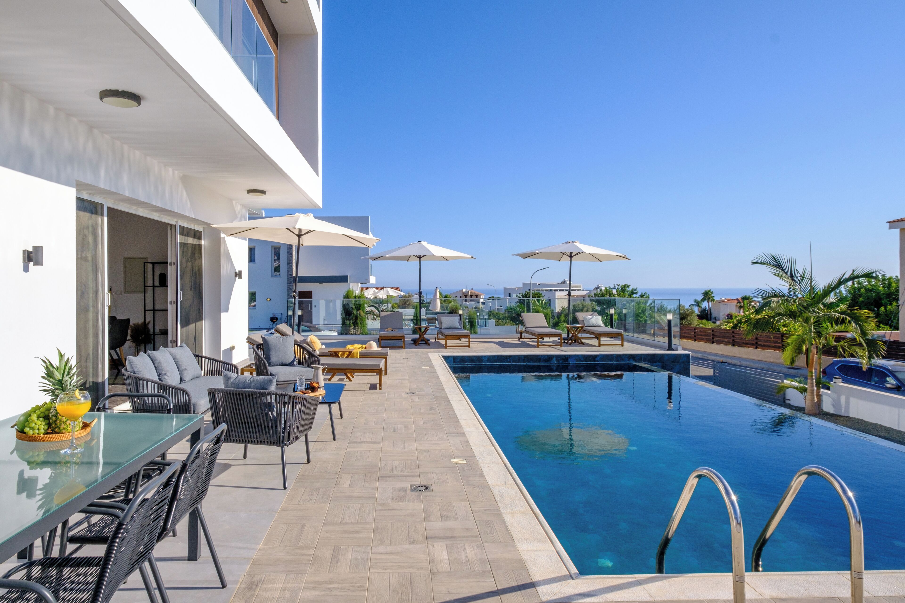 Un espace piscine extérieur moderne, doté de chaises longues et de parasols, d’une table en verre avec des fruits, et offrant une vue sur la mer.