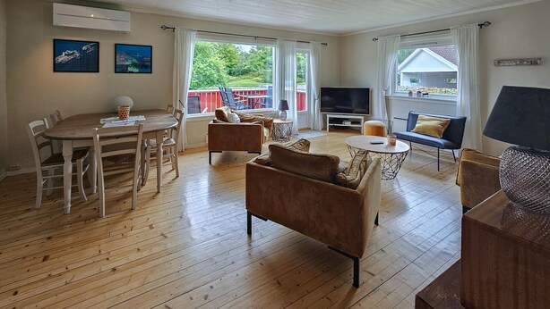A spacious living area with a wooden floor, a dining table with chairs, a sofa, a TV, and a balcony with a view of greenery.