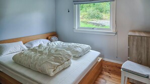 A bedroom with a wooden bed, white bedding, a bedside fan, and a window with a view of greenery.