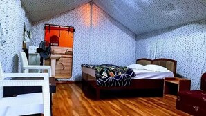 A bedroom with a wooden floor, a bed with a floral bedspread, a desk with a chair, and a small table with a lamp. - Song Valley Resort By Blue Waves (Dehradun)