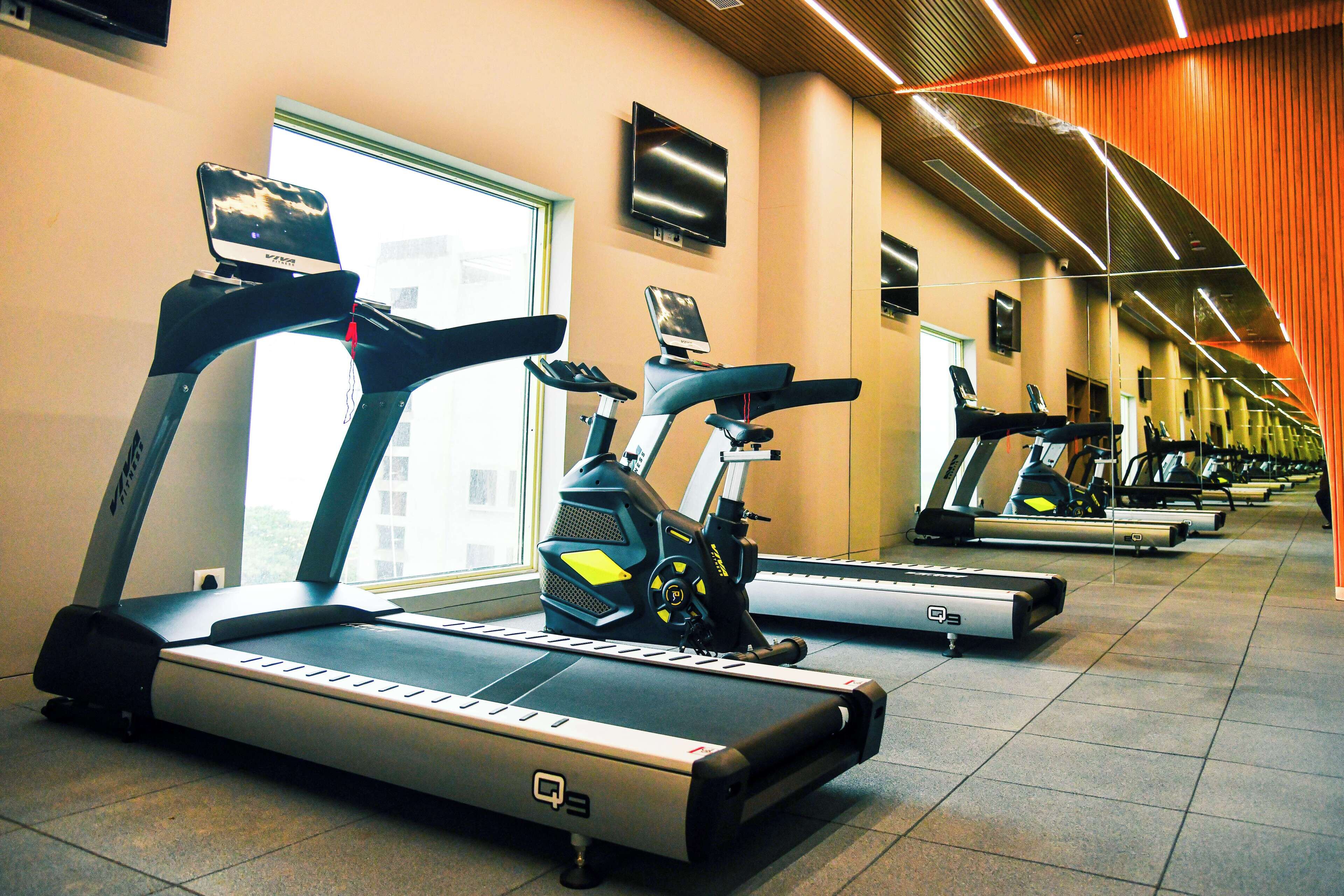 A row of stationary bikes in a gym with a large window and a TV on the wall.