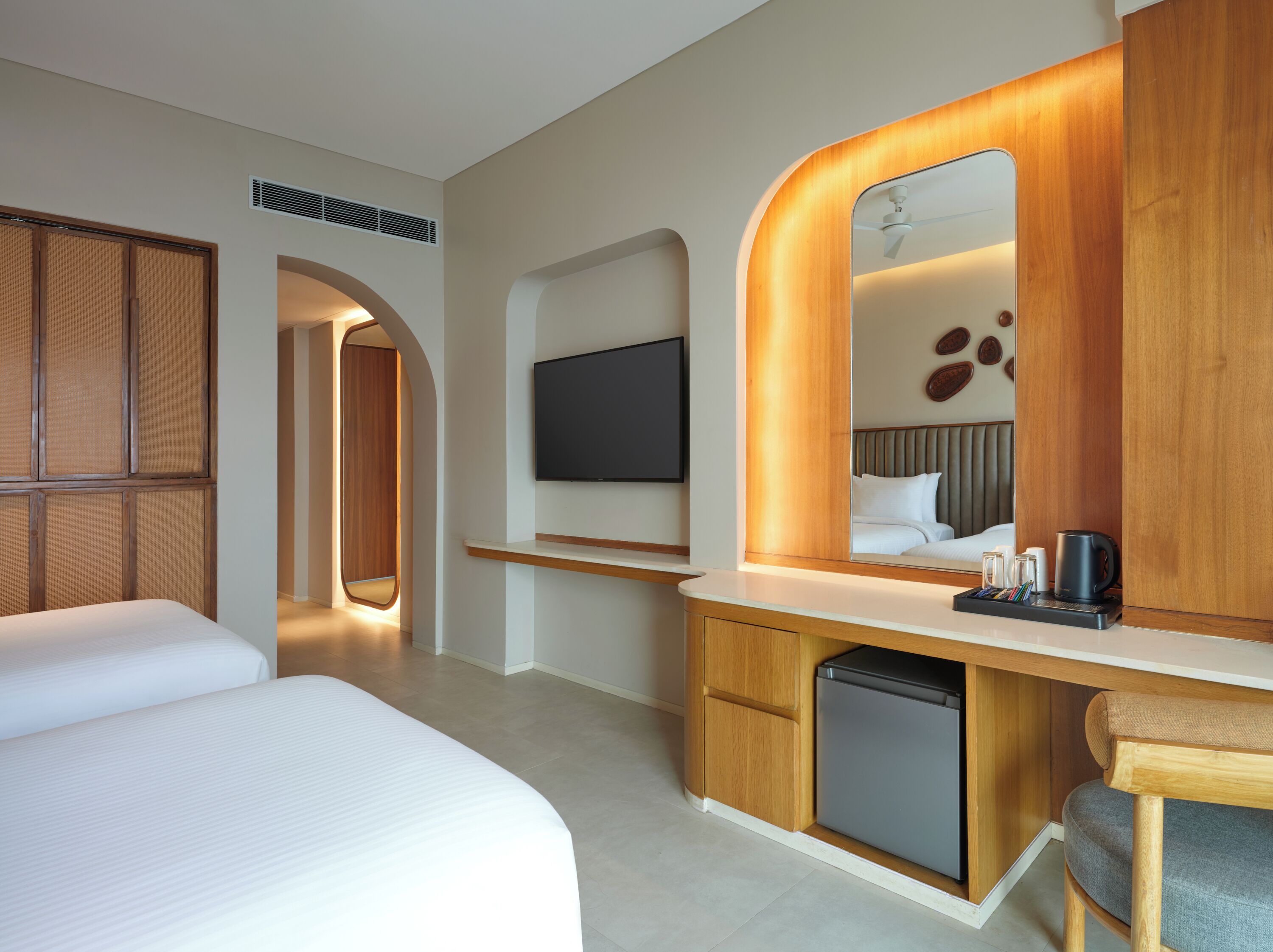 A modern hotel room with a large bed, a small desk with a mirror, a television, and a compact refrigerator.