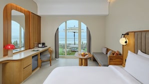 A modern hotel room with a large bed, a desk with a mirror, a small table with fruit, and a view of the ocean. - Swosti Premium Beach Resorts Puri (Brahmagiri)