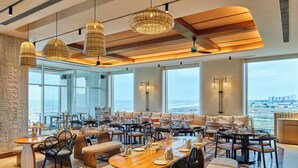 A spacious dining area with multiple tables and chairs, large windows offering a view of the ocean, and hanging wicker lamps. - Swosti Premium Beach Resorts Puri (Brahmagiri)