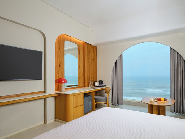 A hotel room with a bed, a desk with a mirror, a television, a view of the ocean, and a small table with a fruit bowl. - Swosti Premium Beach Resorts Puri (Brahmagiri)