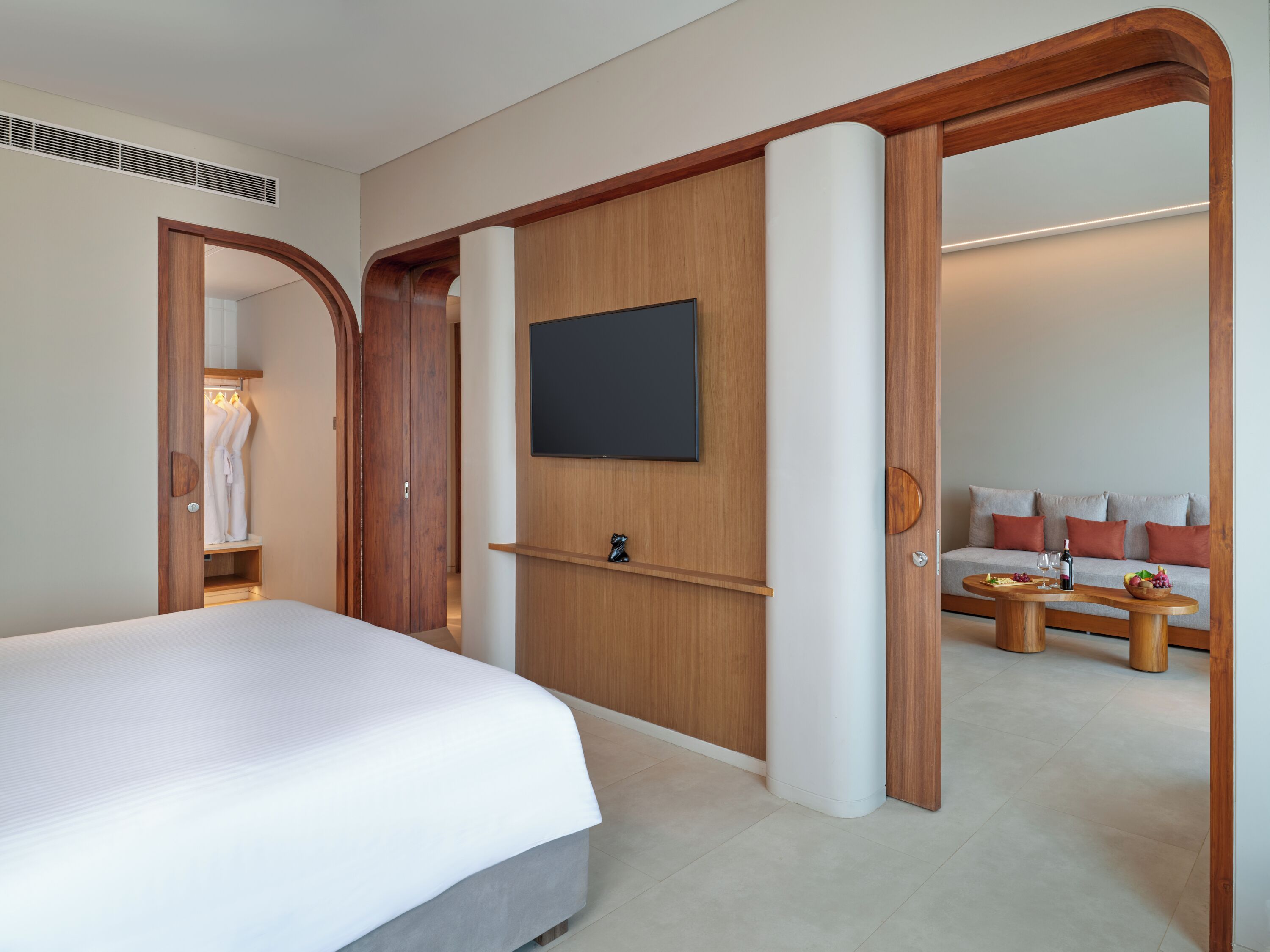 A modern hotel room with a large bed, a flat-screen TV mounted on the wall, a wooden wardrobe, and a seating area with a sofa and a small table.