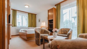A hotel room with a bed, two armchairs, a small table with a vase, a bookshelf, and a window with curtains.