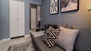 Ein modernes Wohnzimmer mit einem grauen Sofa, gemusterten Kissen und einem geometrischen Teppich. Im Hintergrund sind ein weißer Kleiderschrank und eine Tür zu sehen.