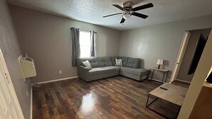 A spacious living room with hardwood flooring, a gray sectional sofa, a ceiling fan, and a small table with a lamp. - Affordable & Cozy Near the Base is set in Lawton. Both free WiFi and parking on-site are accessible at the apartment free of charge! (Lawton)