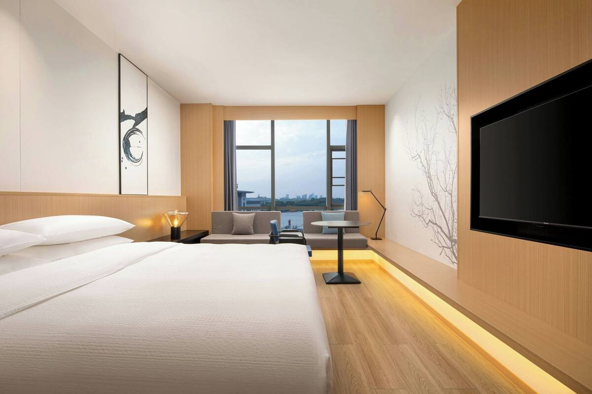 Ein modernes Hotelzimmer mit einem großen Bett, einer Sitzecke mit Tisch, einem Fernseher und Blick auf die Stadt durch große Fenster.