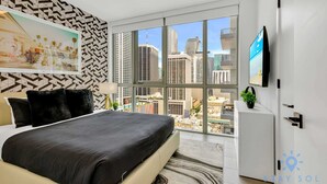 A modern hotel room with a large bed, a city view, a flat-screen TV, and a geometric-patterned wall. - Beautiful Apartment with gym and  amazing Pool (Miami)