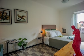 A hotel room with a bed, a desk, a potted plant, and two framed artworks on the wall.