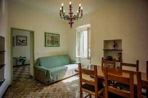 A room with a green sofa, a wooden dining table with chairs, a chandelier, a patterned floor, and a window with curtains.