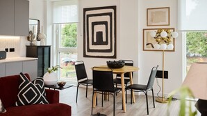A modern living room with a red sofa, black and white patterned cushions, a round wooden table, and black chairs. There is a large abstract painting on the wall, a floor lamp, and a window with a view of greenery outside. - The Tottenham Hale Crib - Trendy 2bdr Flat With Balcony (London)