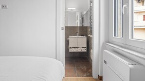 A narrow hallway with a bed on the left, a bathroom with a tiled wall and a mirror, and a window on the right. - Sunny House Gianicolo (roma)
