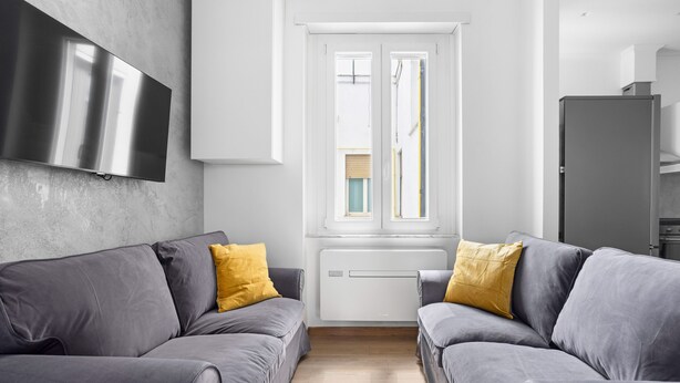 A modern living room with two grey sofas, a flat-screen TV mounted on the wall, and a window with blinds. - Sunny House Gianicolo (roma)