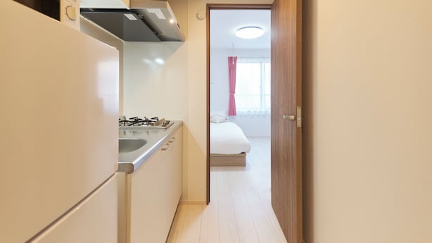 A narrow hallway with a kitchenette on the left, a bed with a pink curtain in the background, and a wooden door on the right. - Sky Up Oshiage by Tranova (Tokyo)